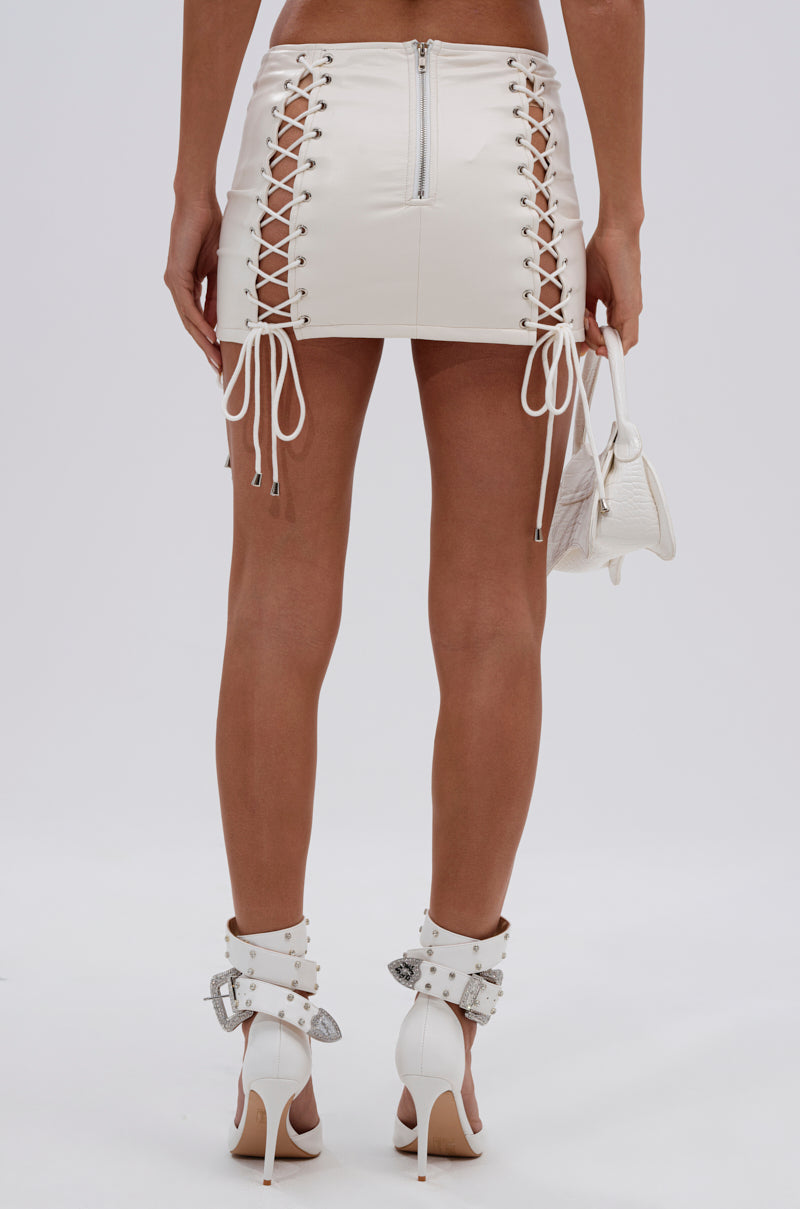 Someone shown from the back against a plain background wears the FESTIVAL GIRLY LACE UP MINI SKIRT, white high-heeled sandals with ankle straps, and holds a small white handbag.