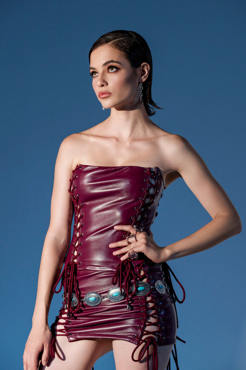 A woman wears the YOU KNOW I WANT YOU BABY LACE UP CORSET, a strapless wine red faux leather dress with lace-up sides and turquoise accents, standing against a blue background and looking off to the side with her hand on her hip.