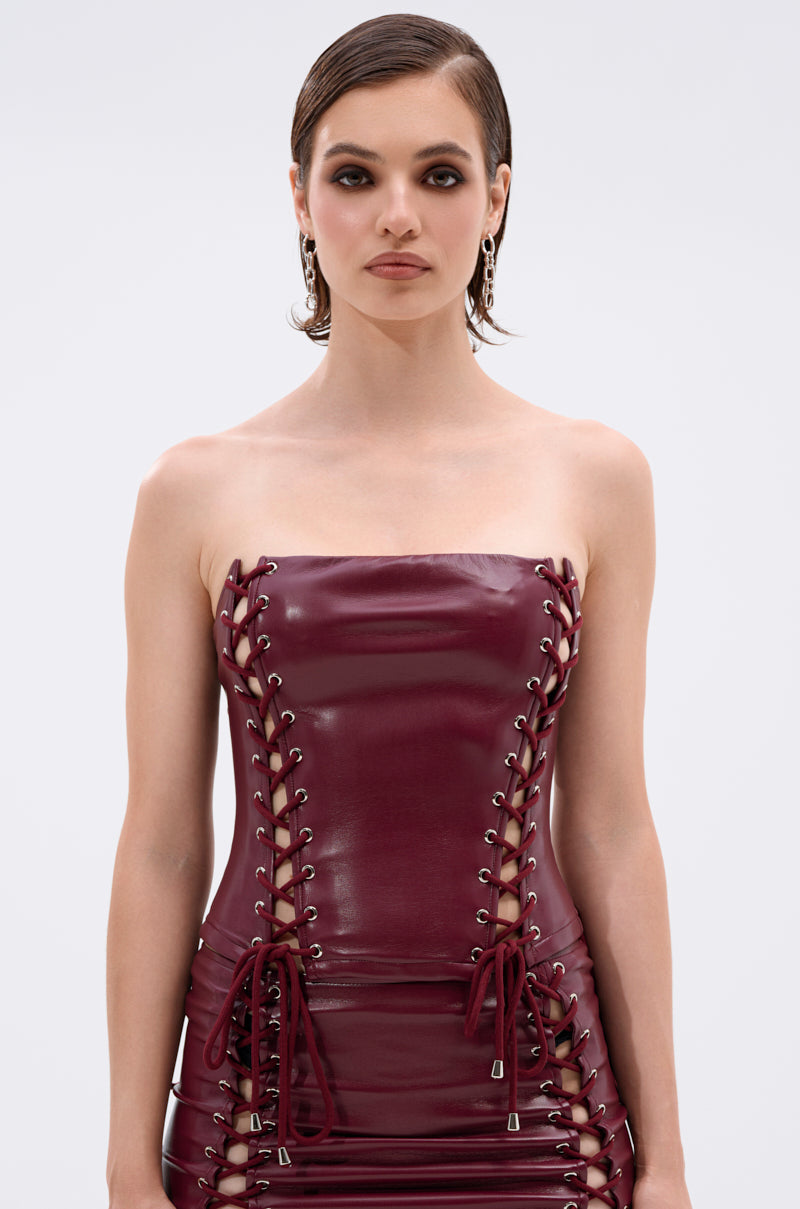 A woman with short, slicked-back hair wears the YOU KNOW I WANT YOU BABY LACE UP CORSET, a wine-red strapless top with front bows, styled like latex and laced up, against a plain light background.