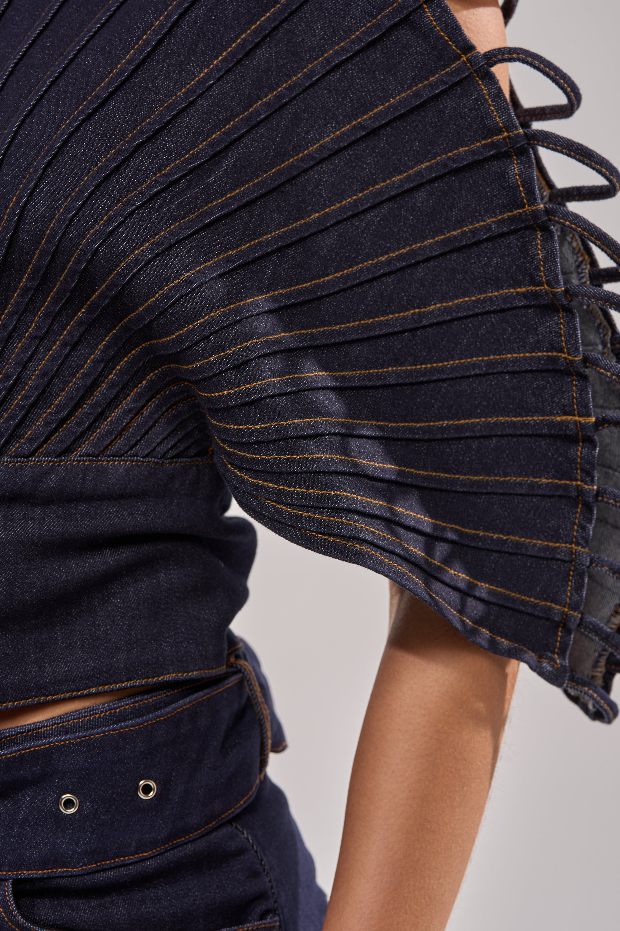 Close-up of a person wearing the DENIM ON DENIM BLOUSE IN DARK BLUE DENIM, featuring sculptural pleated fan sleeves with visible stitching and a matching high-waisted belt with metal grommets.