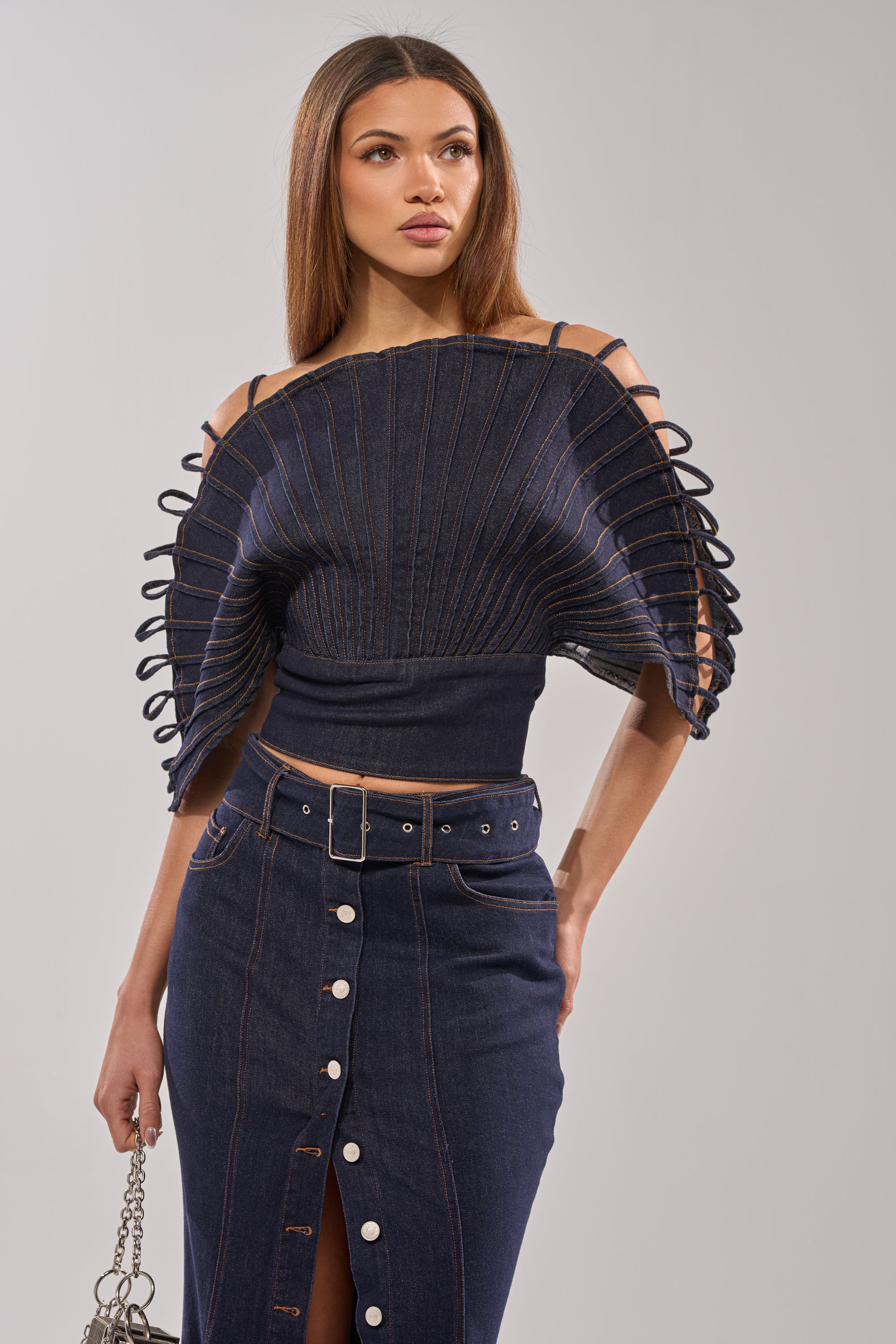 A woman makes a statement in the DENIM ON DENIM BLOUSE IN DARK BLUE DENIM with sculptural sleeves and circular details, styled with a high-waisted button-front skirt and silver chain bag.