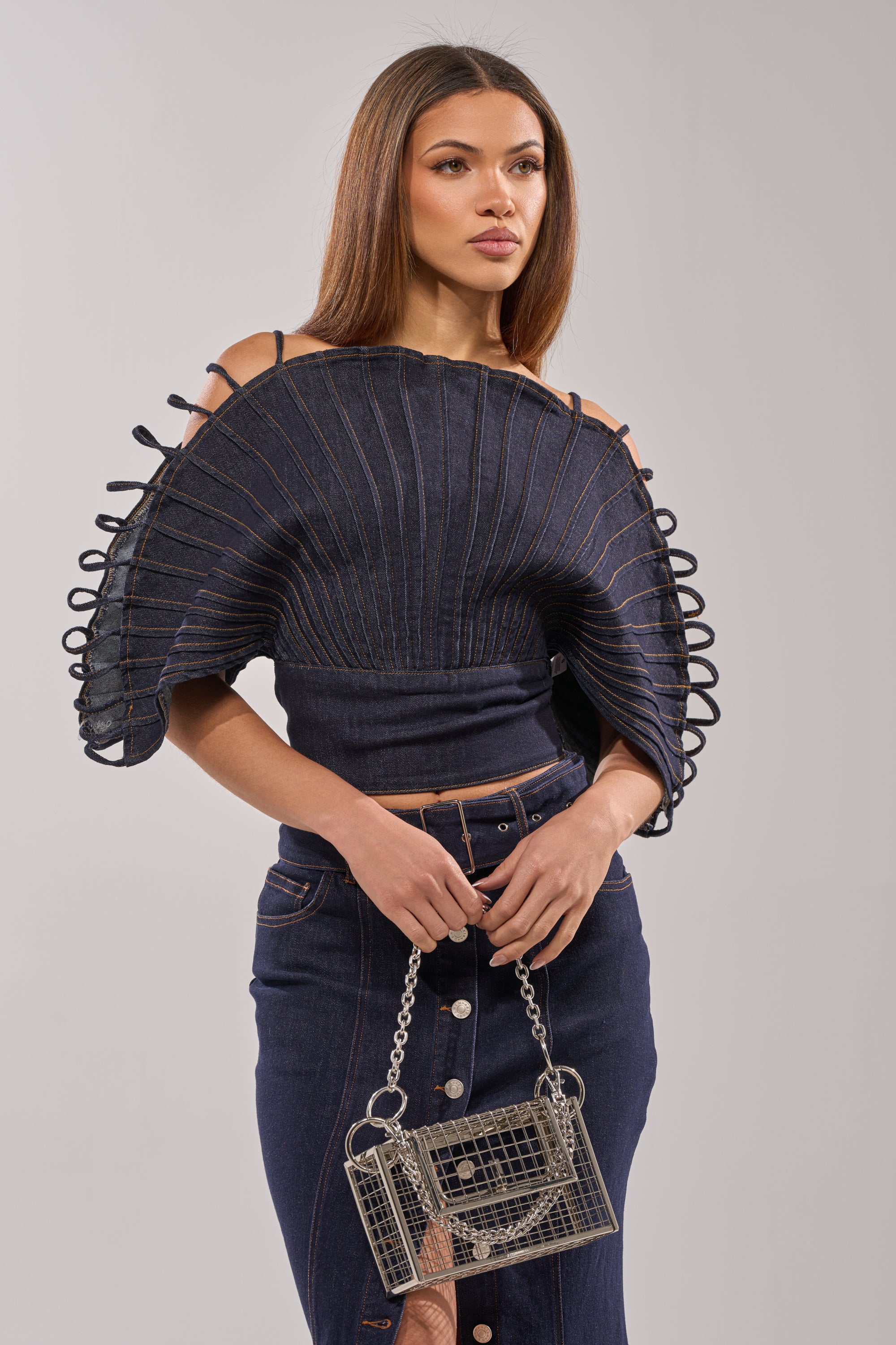 A woman wears the DENIM ON DENIM BLOUSE IN DARK BLUE DENIM featuring structured pleats and edge loops, paired with a matching skirt. She carries a small boxy chain purse and has straight brown hair.