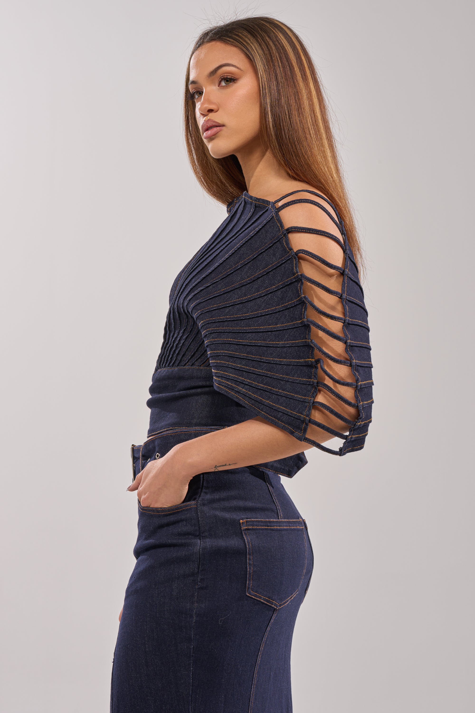 A woman in a dark denim outfit stands sideways, wearing the DENIM ON DENIM BLOUSE IN DARK BLUE DENIM with sculptural sleeves and high-waisted jeans, against a plain light gray background.