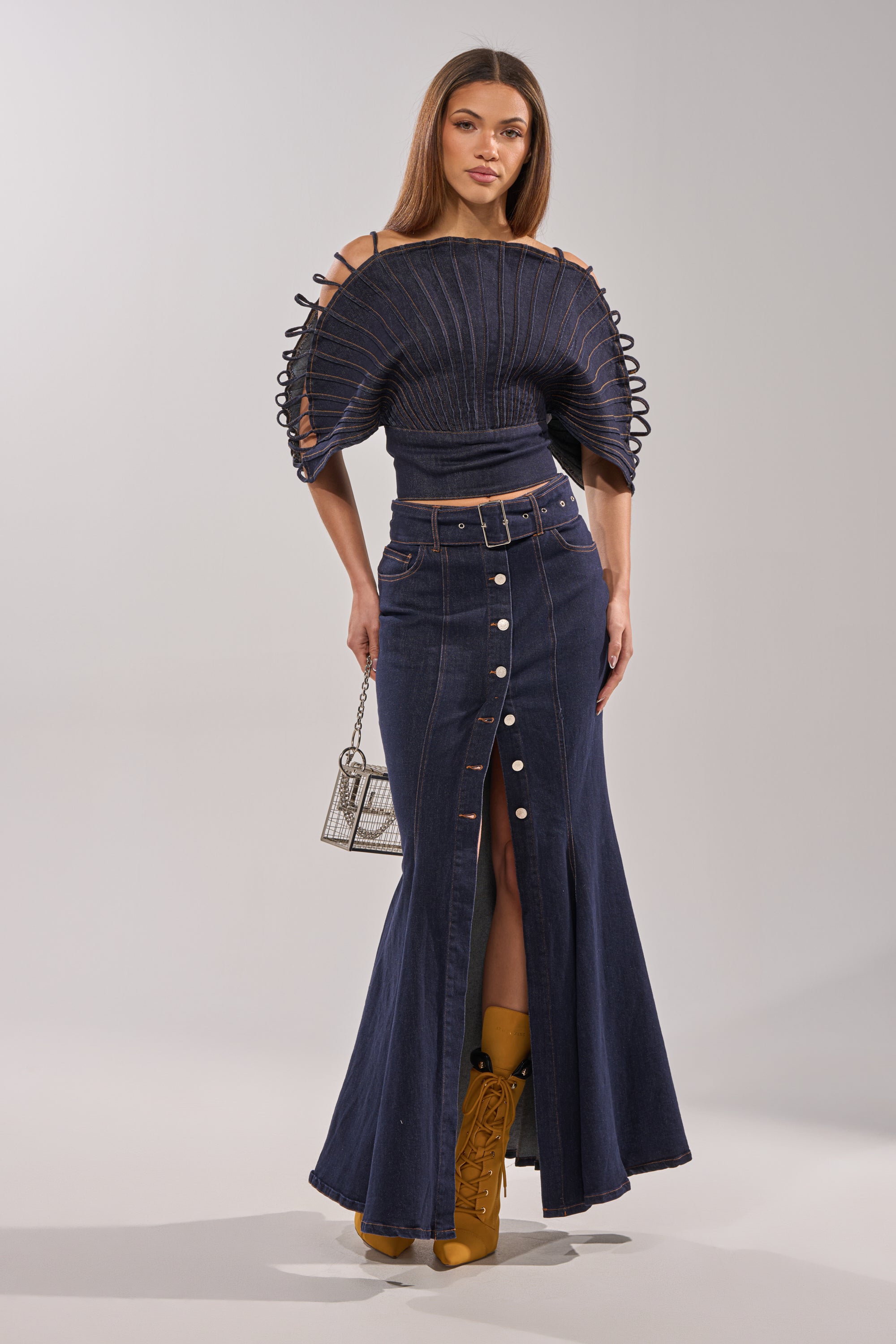 A woman makes a fashion statement in the DENIM ON DENIM BLOUSE IN DARK BLUE DENIM, styled with a long, high-waisted denim skirt with front buttons and a slit. She completes the look with a small handbag and tan boots.