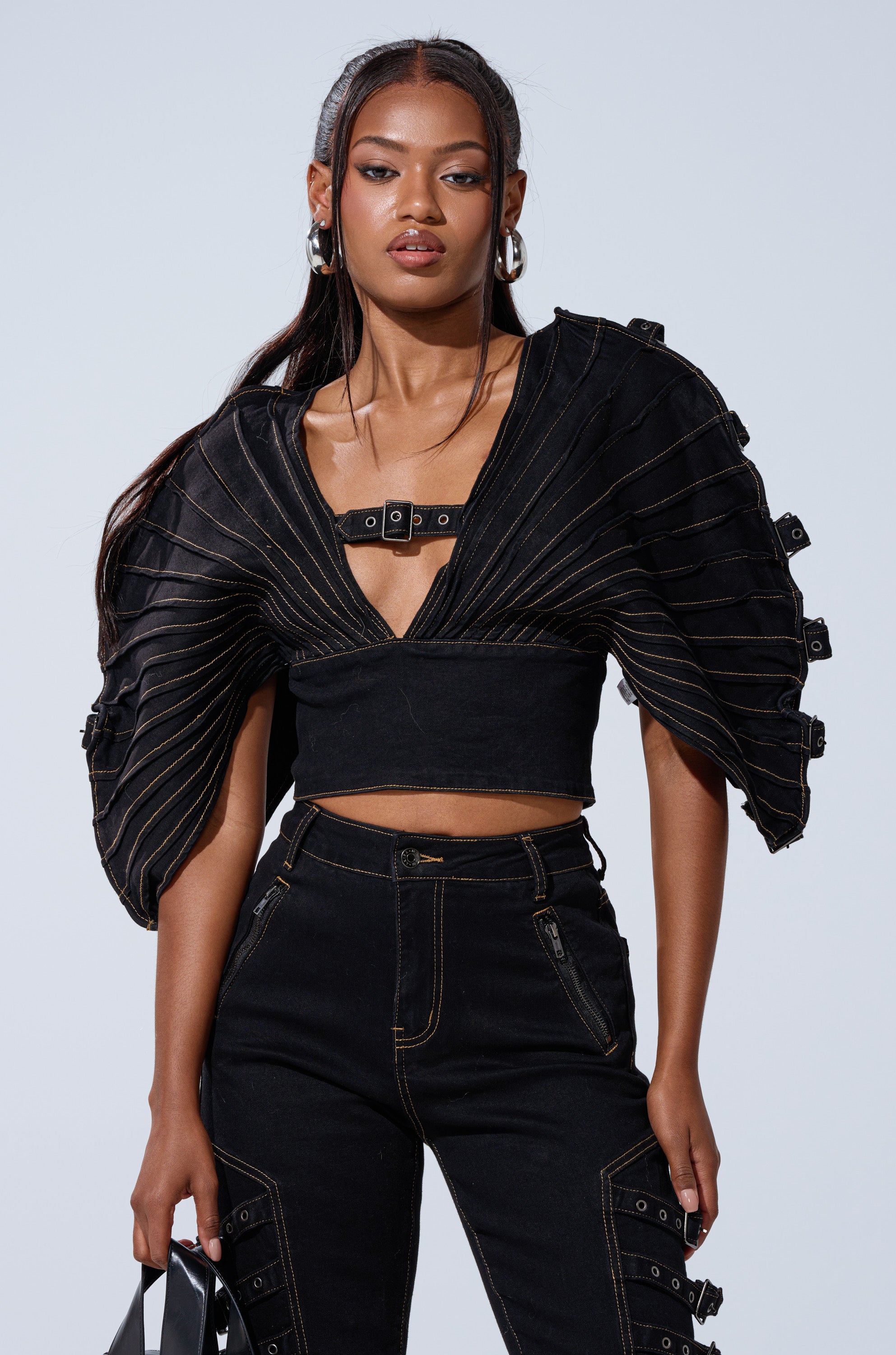 A woman wears the BUCKLE UP DENIM BLOUSE with winged sleeves and a front buckle, styled with high-waisted black pants featuring exposed stitching and zippers. She carries a black bag and has long, straight hair.