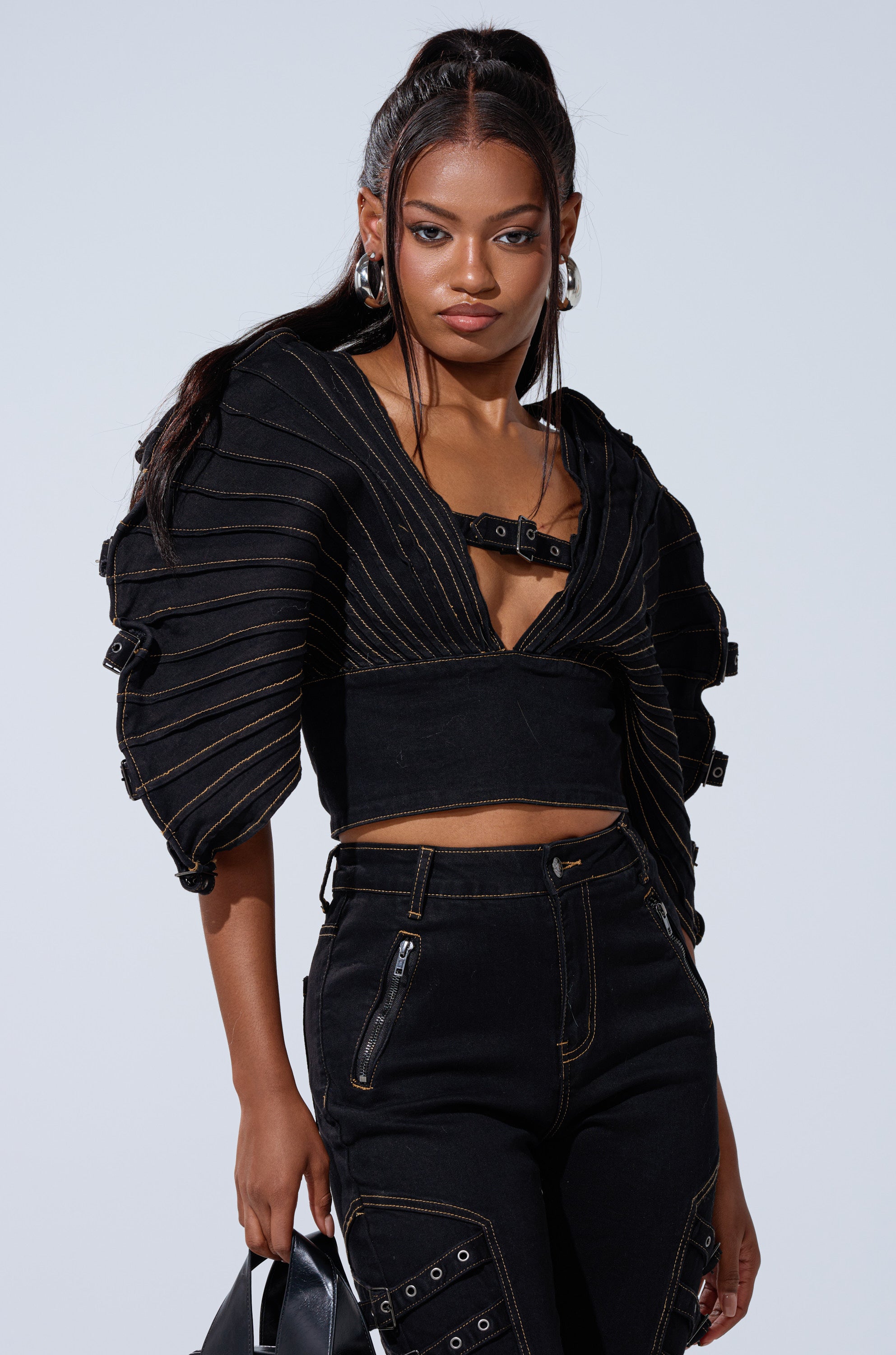 A woman with long, wavy dark hair wears the BUCKLE UP DENIM BLOUSE, featuring puffed pleated sleeves and a deep V-neckline. She pairs it with high-waisted black pants with zipper and buckle details, and holds a black handbag.