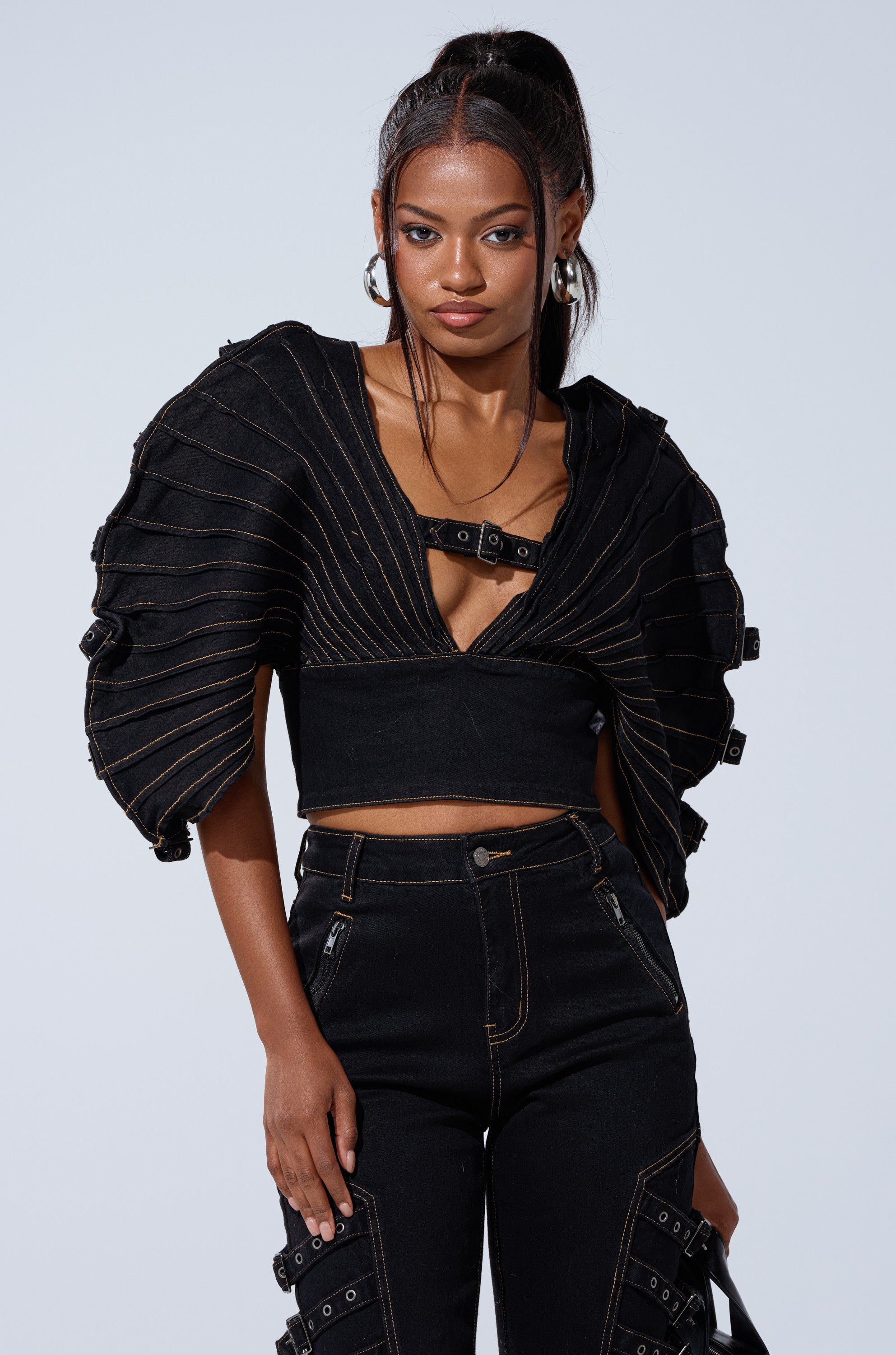 A woman models the BUCKLE UP DENIM BLOUSE—a black denim top with bold, structured sleeves and decorative stitching—standing confidently against a plain light background, her hair in a high ponytail and wearing minimal jewelry.