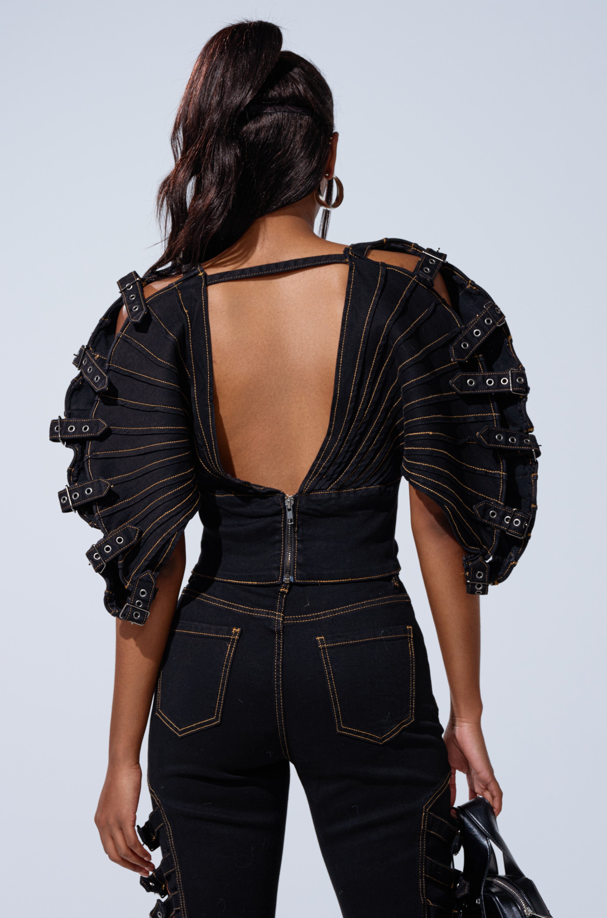 A woman with long dark hair in a ponytail models the BUCKLE UP DENIM BLOUSE—a black open-back top featuring dramatic ruffled sleeves with buckles and matching high-waisted pants—against a plain light background.