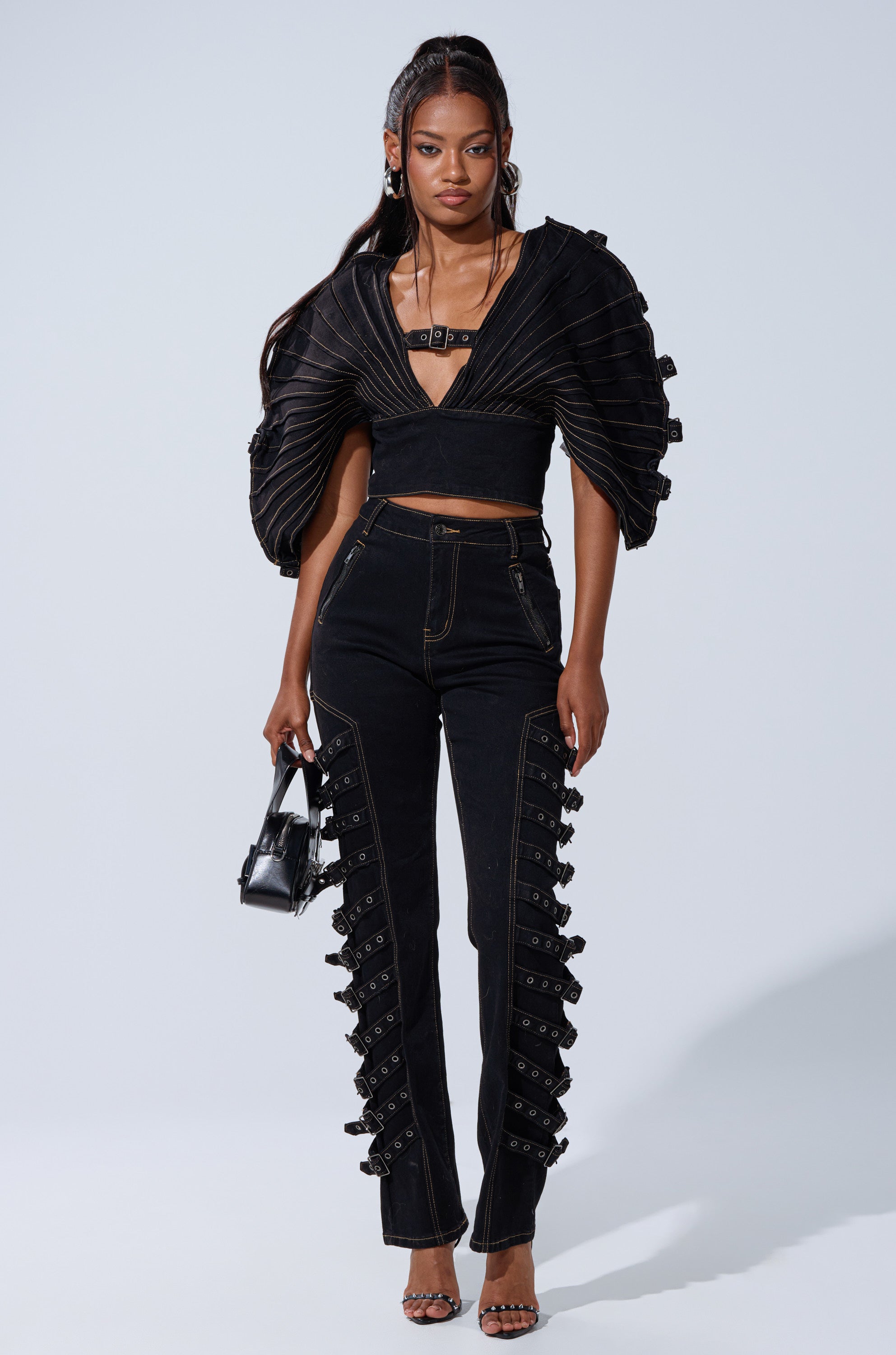 A woman stands against a plain background wearing the BUCKLE UP DENIM BLOUSE with voluminous sleeves, paired with matching high-waisted pants featuring buckle details. She accessorizes with a small black handbag and heeled sandals.