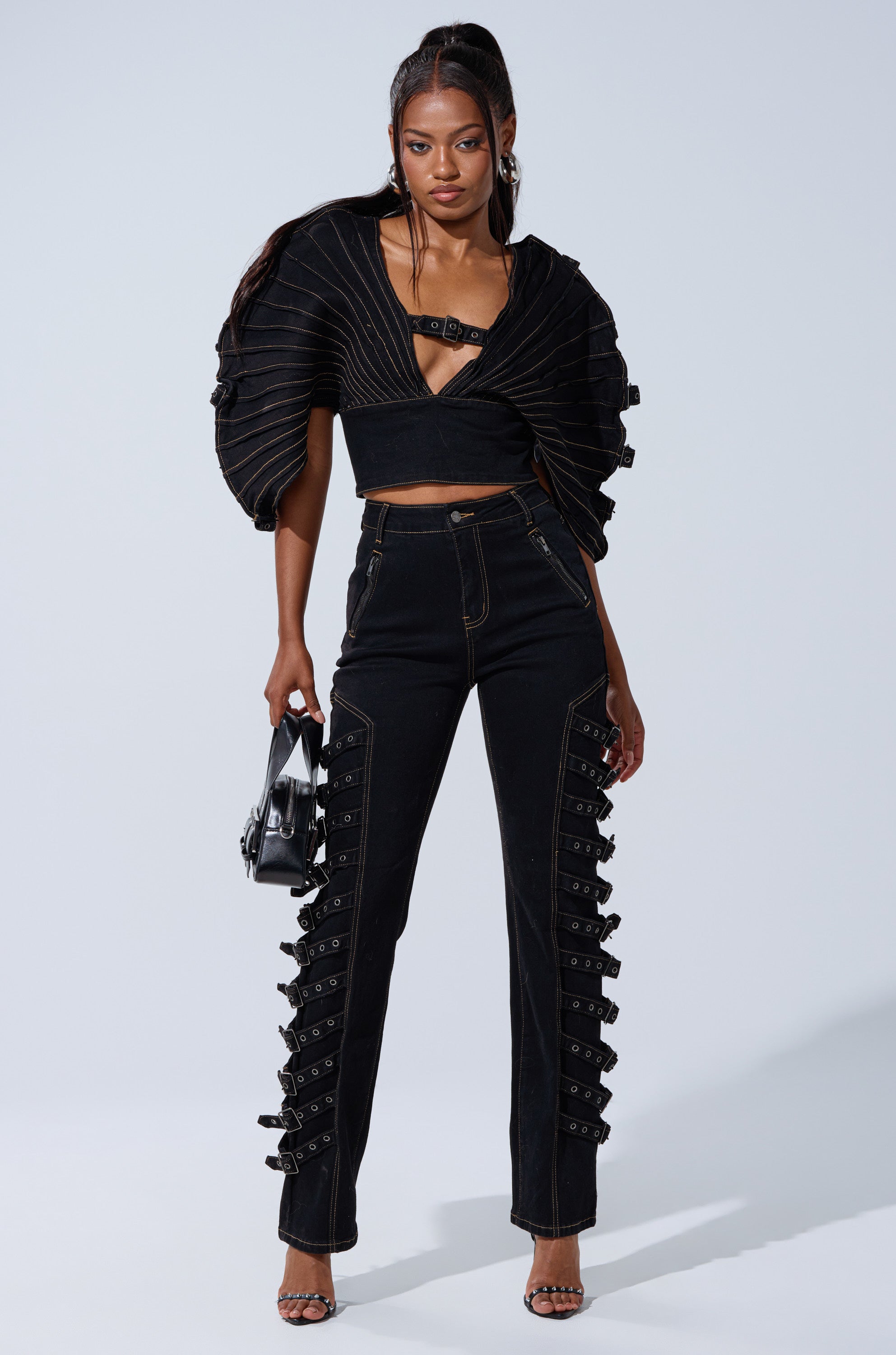 A woman wears the BUCKLE UP DENIM BLOUSE with bold shoulders, paired with high-waisted pants featuring decorative buckles, strappy heels, and a small black handbag as she stands confidently against a plain background.