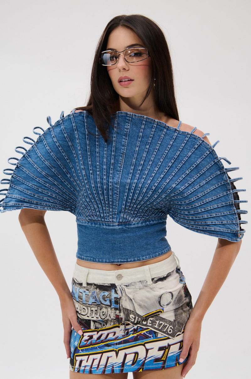 A woman wears tinted glasses and the DENIM ON DENIM BLOUSE—a pleated denim top with radiating structured strips—paired with a printed denim skirt, standing against a plain white background with hands on her hips.