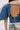 A person wears the DENIM ON DENIM BLOUSE, featuring a blue dramatic pleated silhouette and open back, paired with light patchwork pants with bold text and graphic prints. Only their torso is visible.