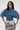 A woman wears tinted glasses and the DENIM ON DENIM BLOUSE—a pleated denim top with radiating structured strips—paired with a printed denim skirt, standing against a plain white background with hands on her hips.