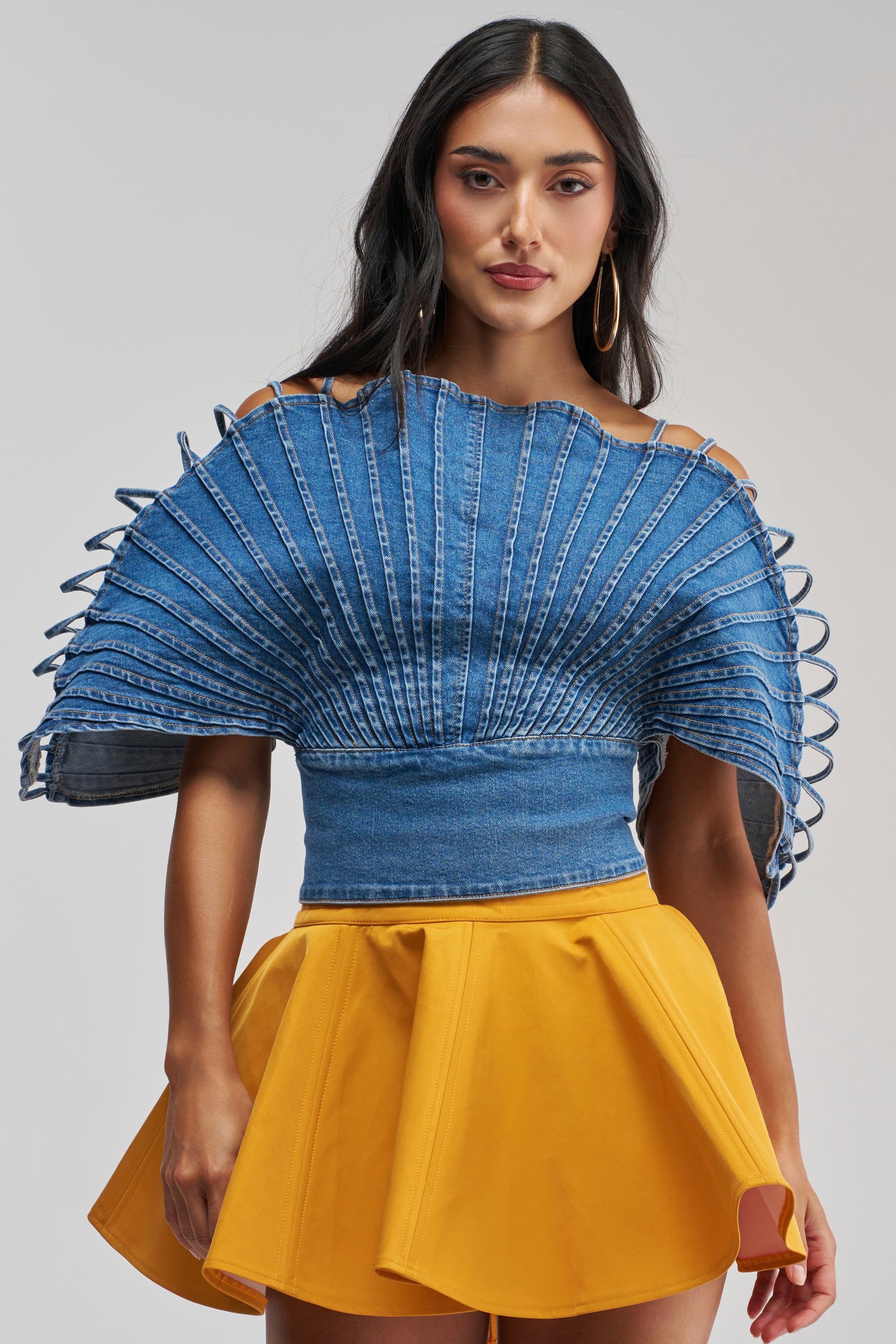 A woman with long dark hair and gold hoop earrings stands against a plain background, wearing the DENIM ON DENIM BLOUSE—a blue pleated denim top featuring unique looped details—paired with a bright yellow skirt for the perfect date night look.