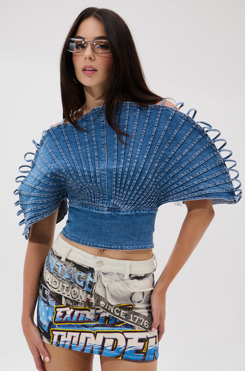A woman with long dark hair wears the DENIM ON DENIM BLOUSE, featuring a pleated silhouette and sculpted details, paired with high-waisted graphic print shorts and stylish rectangular glasses.