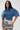 A woman with long dark hair wears the DENIM ON DENIM BLOUSE, featuring a pleated silhouette and sculpted details, paired with high-waisted graphic print shorts and stylish rectangular glasses.