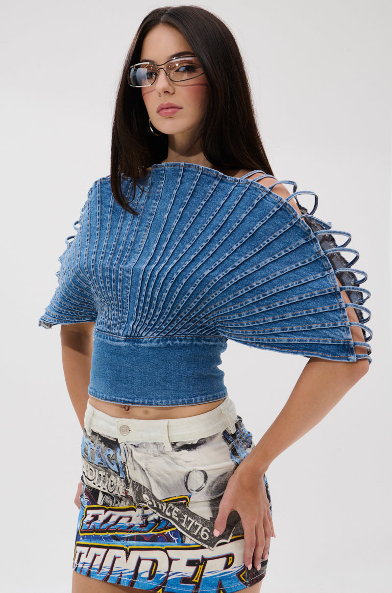 A woman in large glasses and the DENIM ON DENIM BLOUSE with a pleated silhouette and lace-up sides, paired with a printed mini skirt, stands against a plain white background.