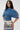 A woman in large glasses and the DENIM ON DENIM BLOUSE with a pleated silhouette and lace-up sides, paired with a printed mini skirt, stands against a plain white background.