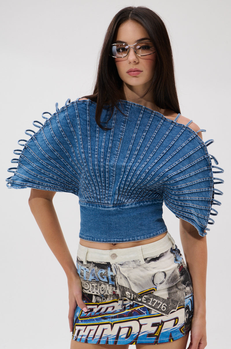 A woman in the DENIM ON DENIM BLOUSE with looped edges, fan sleeves, and an open back, styled with a colorful graphic mini skirt and sunglasses, stands against a plain white background.