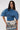 A woman in the DENIM ON DENIM BLOUSE with looped edges, fan sleeves, and an open back, styled with a colorful graphic mini skirt and sunglasses, stands against a plain white background.