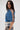 A woman with long dark hair and clear glasses models the DENIM ON DENIM BLOUSE with open crisscrossed sleeves and back, paired with high-waisted printed pants. She stands sideways against a plain white background.