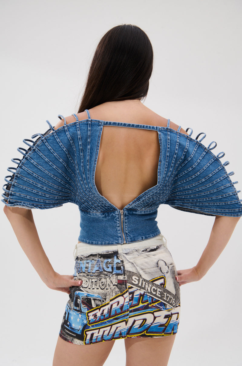 A woman faces away from the camera, wearing the DENIM ON DENIM BLOUSE with dramatic pleated sleeves and an open back, paired with high-waisted shorts adorned with colorful graphics and text.