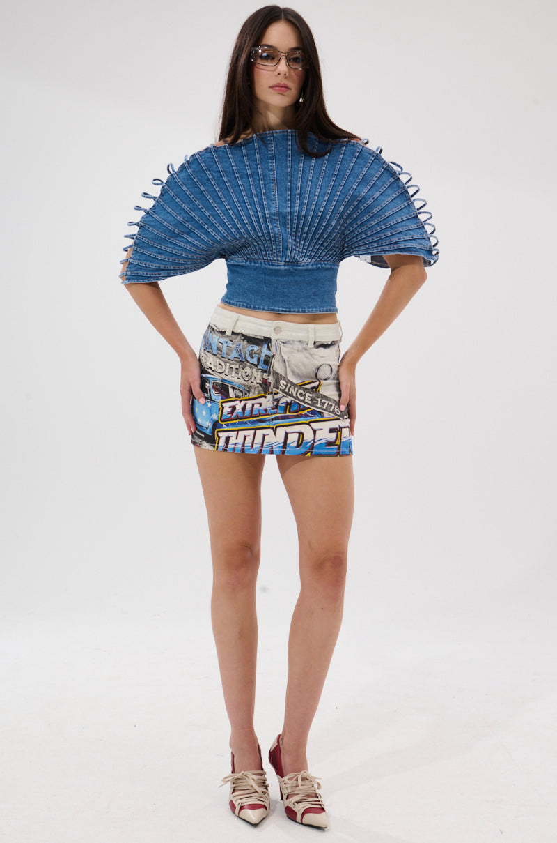 A woman with long dark hair and sunglasses wears the DENIM ON DENIM BLOUSE with wide, structured sleeves, paired with a graphic denim skirt and pointed strap shoes. The background is plain white.