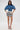 A woman with long dark hair and sunglasses wears the DENIM ON DENIM BLOUSE with wide, structured sleeves, paired with a graphic denim skirt and pointed strap shoes. The background is plain white.