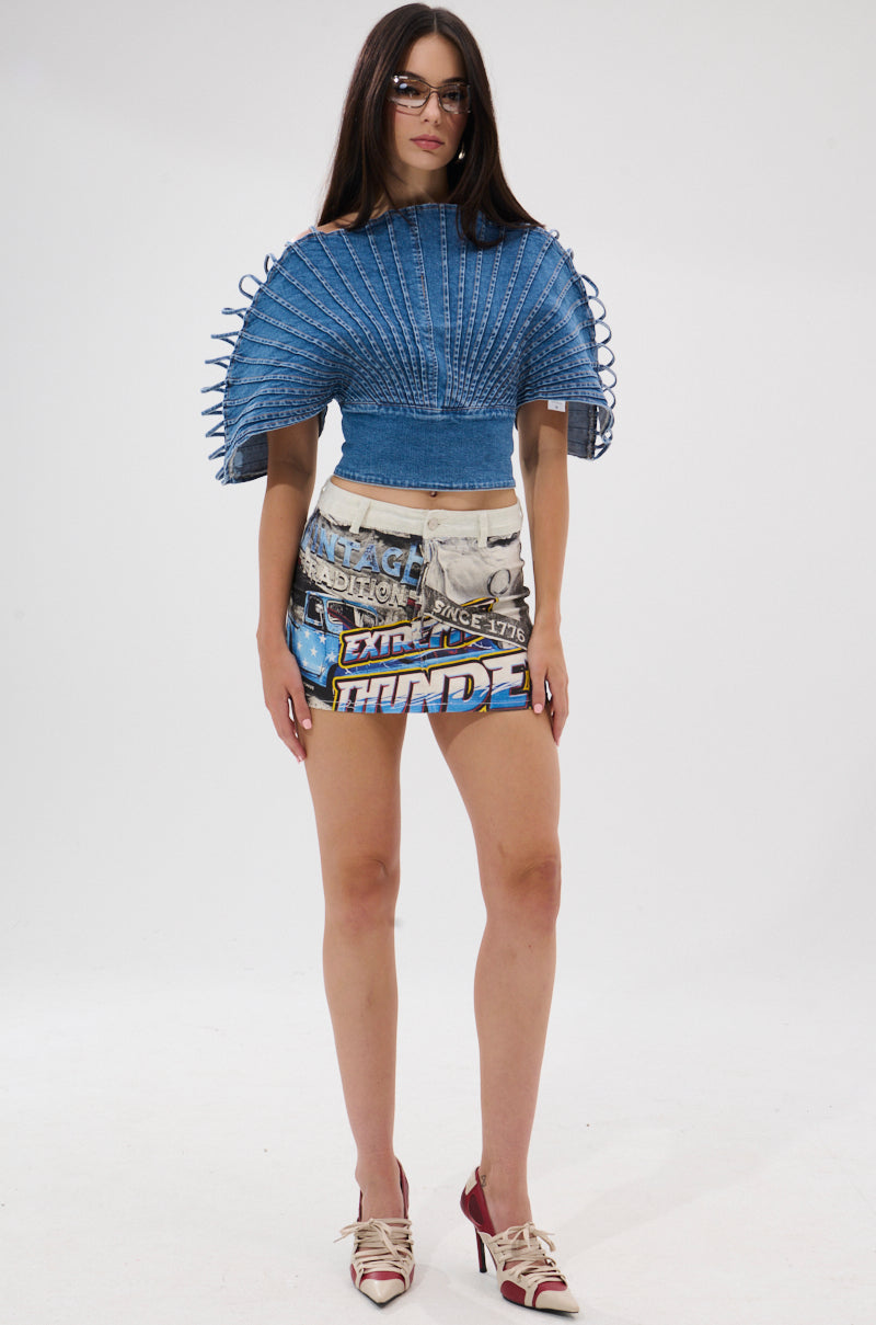 A woman with long brown hair, glasses, and high heels faces forward, wearing the DENIM ON DENIM BLOUSE in blue with pleated details and laced sides, paired with graphic text mini shorts. The background is plain white.
