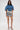 A woman with long brown hair, glasses, and high heels faces forward, wearing the DENIM ON DENIM BLOUSE in blue with pleated details and laced sides, paired with graphic text mini shorts. The background is plain white.