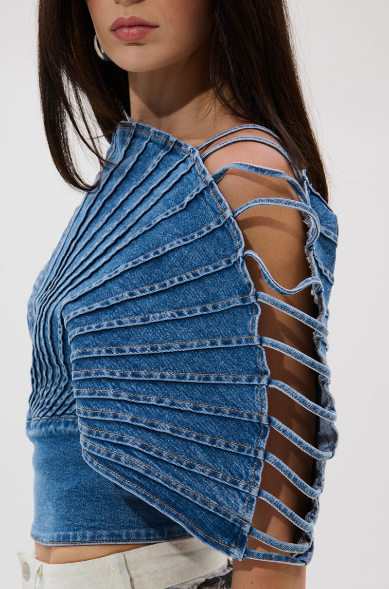 A woman wears the DENIM ON DENIM BLOUSE, a unique blue denim top with radiating pleats and open lattice sleeves, paired with high-waisted light pants. The blouse features a striking pleated silhouette; only her lower face is visible.