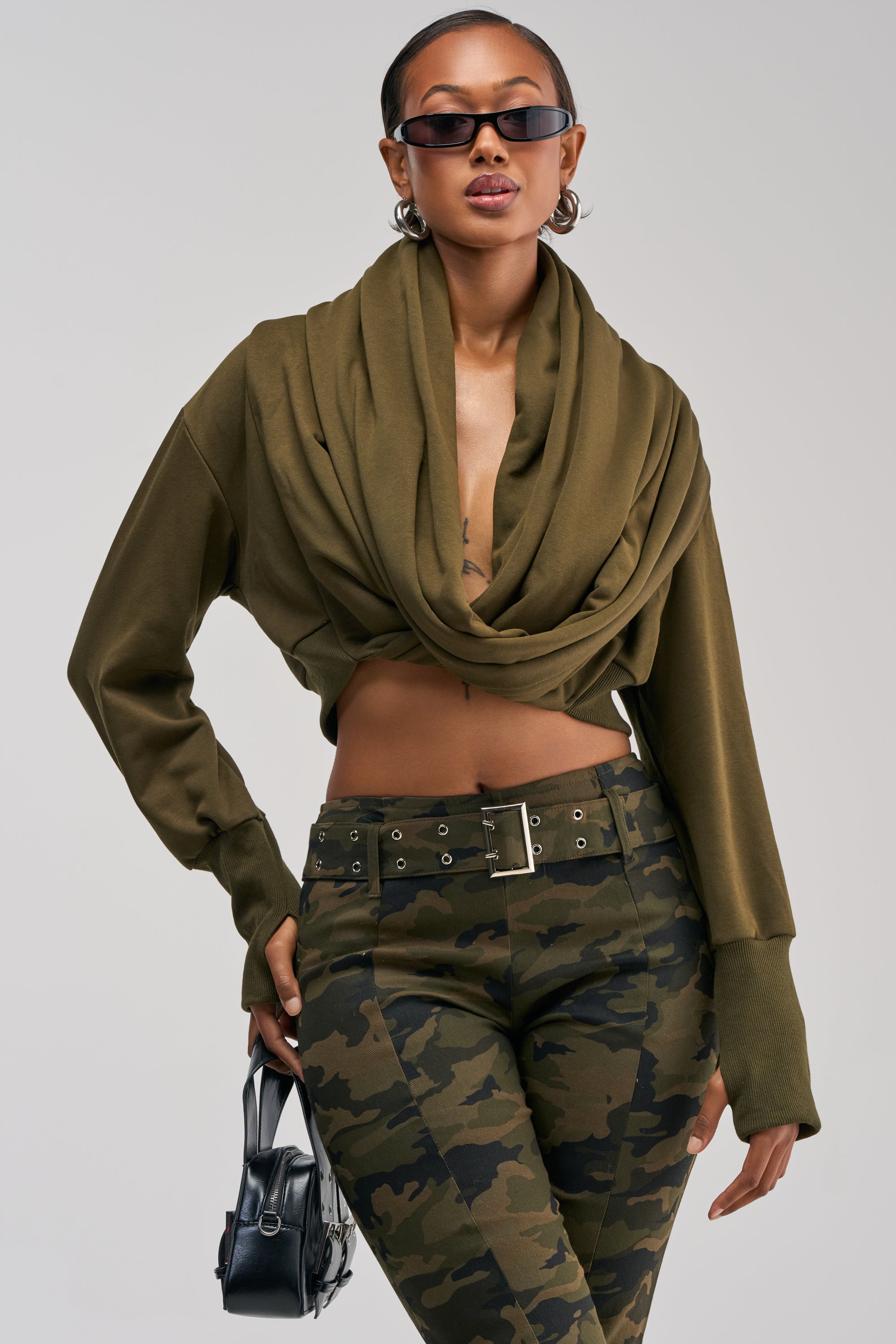 A woman wearing sunglasses and the JUST TOO COOL FOR YOU SWEATSHIRT IN OLIVE, styled as streetwear with camouflage pants, a wide belt, and a small black handbag, poses confidently against a plain background.