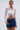 A woman wears the GIMME MORE LACE ZIP-UP TOP in white with a blue denim mini skirt, hoop earrings, a nose ring, and a belly button piercing. She holds a white bag against a plain light background.