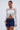 A woman wearing the GIMME MORE LACE ZIP-UP TOP in white with long sleeves and a blue denim mini skirt stands against a plain light background, holding a small white handbag and looking slightly to the side.