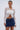 A woman stands against a plain light background wearing the GIMME MORE LACE ZIP-UP TOP— a white floral lace long sleeve zip-up—paired with a blue denim mini skirt, hoop earrings, tied-back hair, and holding a white handbag.