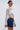 A woman wears the GIMME MORE LACE ZIP-UP TOP in white with long sleeves, paired with a dark denim mini skirt and holding a small white bag, posing sideways against a plain light background.