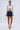 A woman stands against a plain background wearing the GIMME MORE LACE ZIP-UP TOP, a dark blue denim mini skirt, white high-heeled sandals, hoop earrings, and holding a small white handbag.