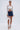A woman stands against a plain background wearing the GIMME MORE LACE ZIP-UP TOP, a dark blue denim mini skirt, white heeled ankle-strap sandals, hoop earrings, and holding a small white handbag.