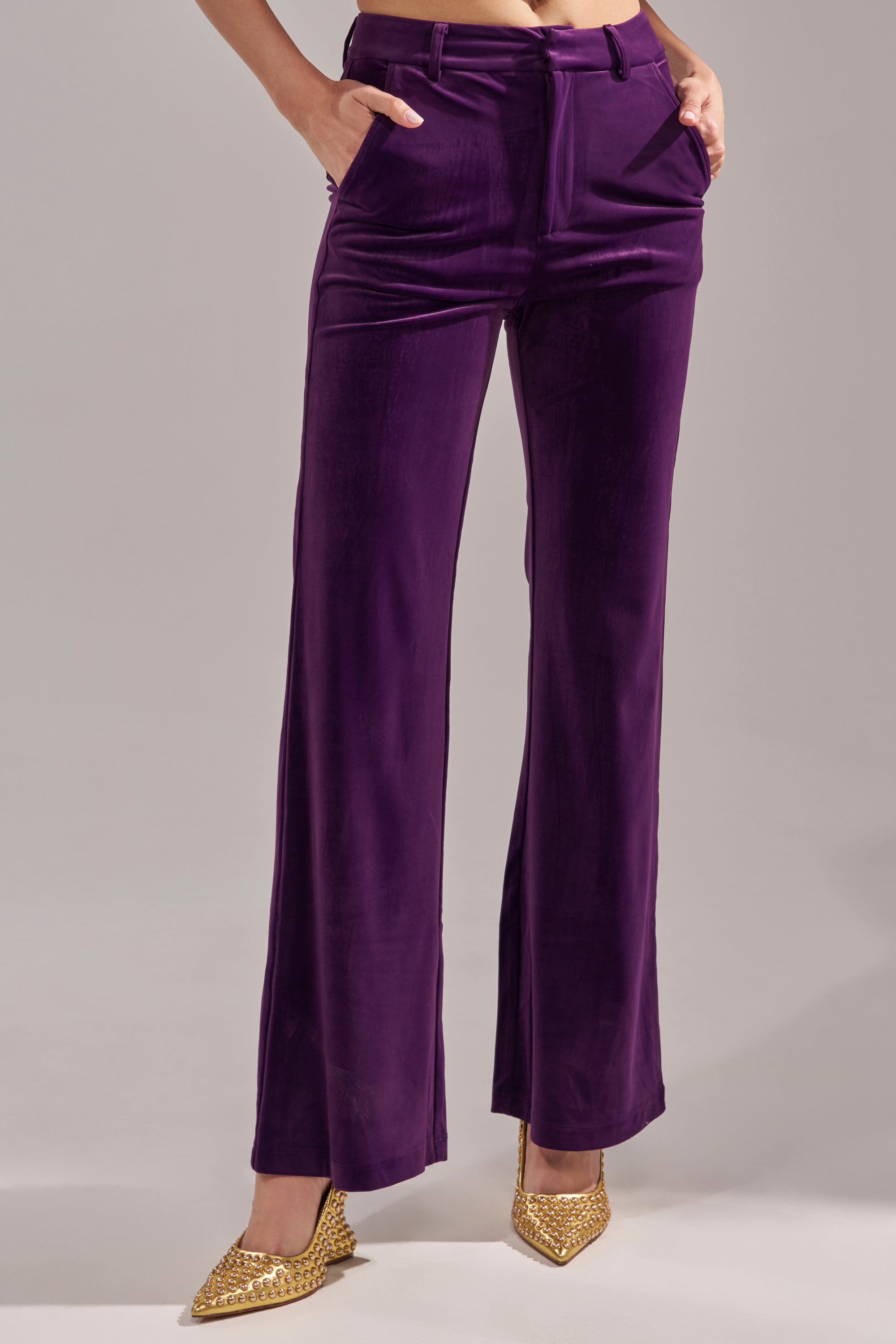 A person models the I ADORE YOU TROUSER in high-rise eggplant purple velvet, hands in pockets, paired with pointed gold shoes, standing against a plain gray background.
