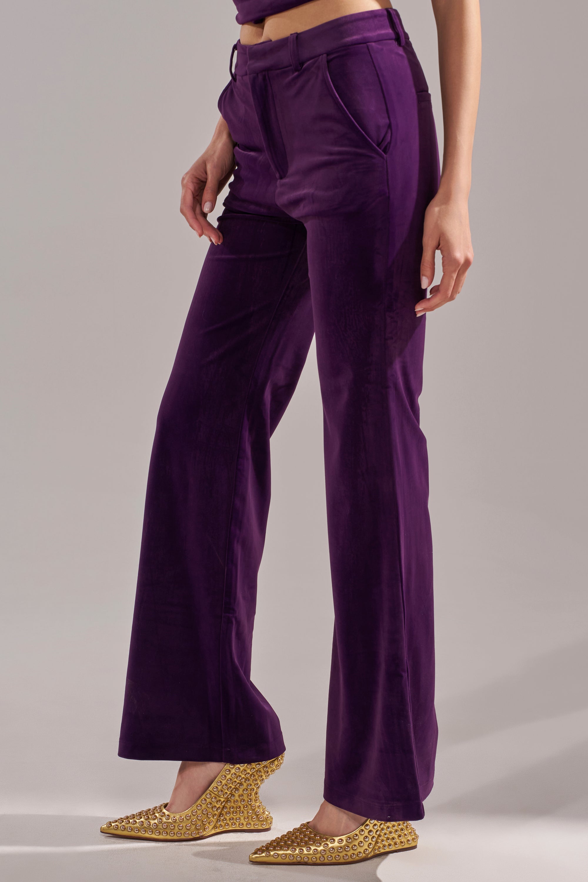 Someone stands against a neutral background wearing the I ADORE YOU TROUSER in eggplant purple velvet and gold textured pointed-toe shoes.
