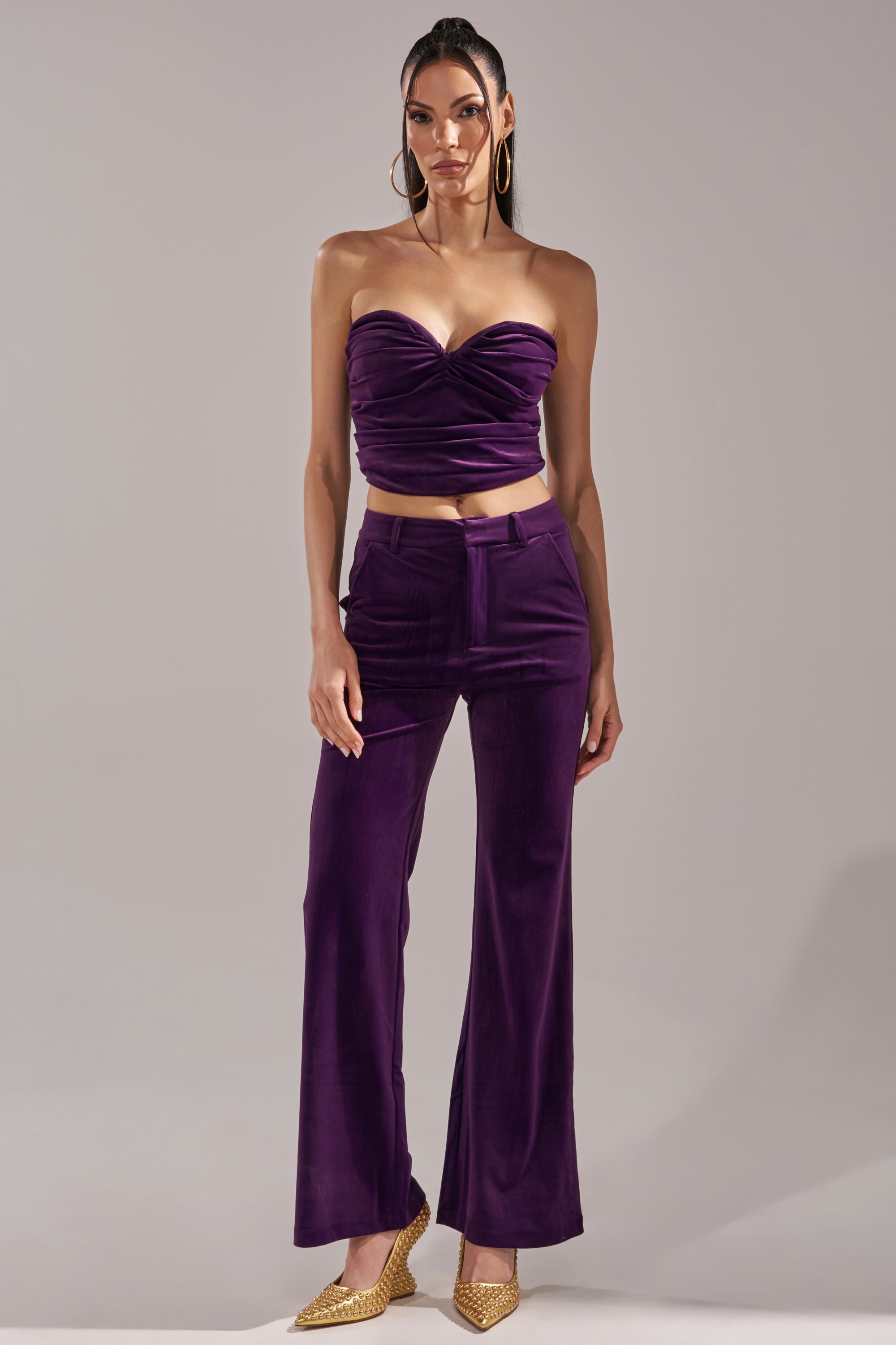 A woman poses against a plain light background in the I ADORE YOU TROUSER, paired with a strapless, ruched eggplant purple velvet crop top. She accessorizes with long hair, large hoop earrings, and gold pointed-toe shoes.