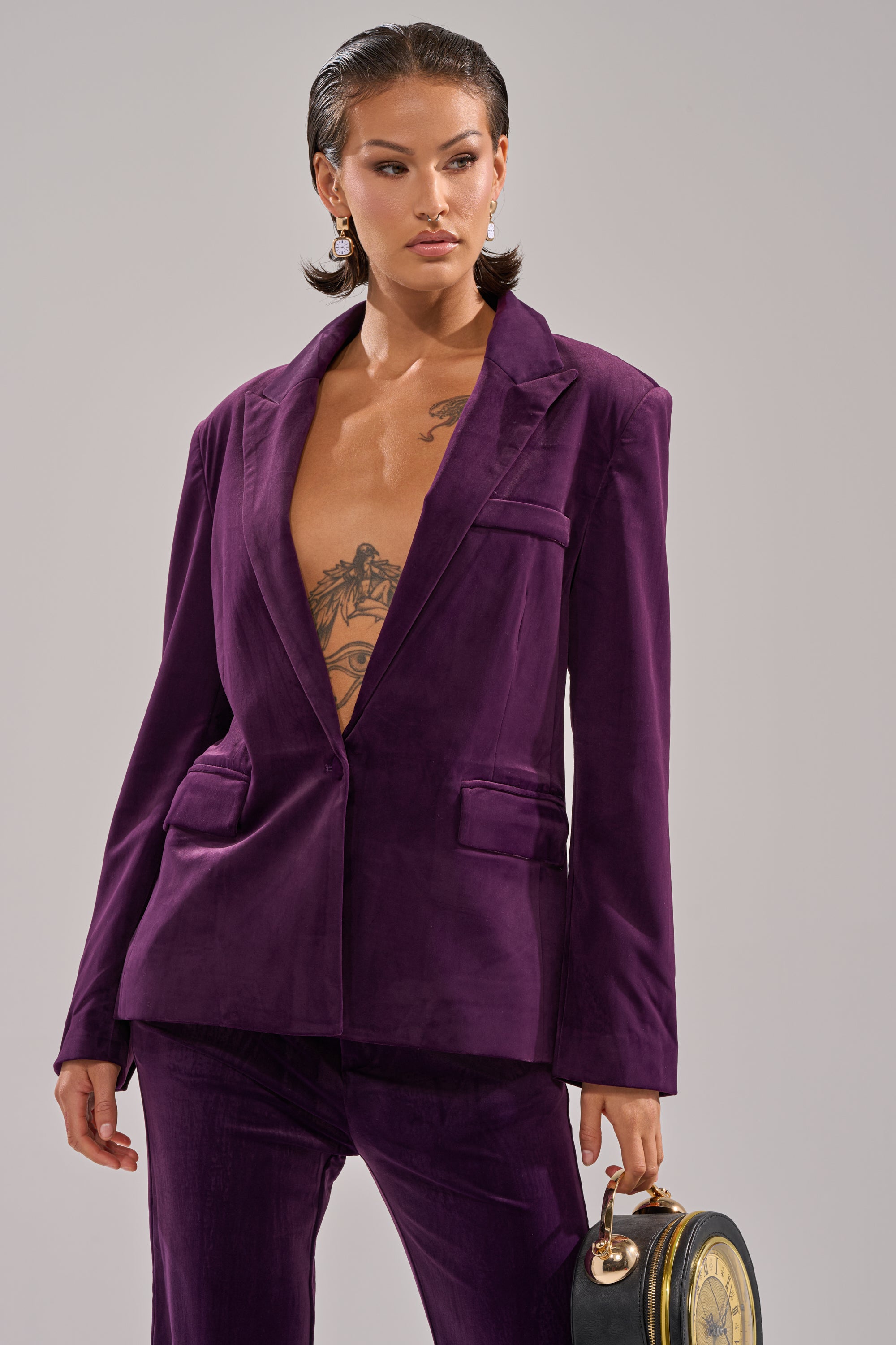 A person with slicked-back hair wears the I ADORE YOU BLAZER in plum purple velvet with matching pants, no shirt underneath to reveal a chest tattoo. They accessorize with a round black handbag and small dangling earrings.