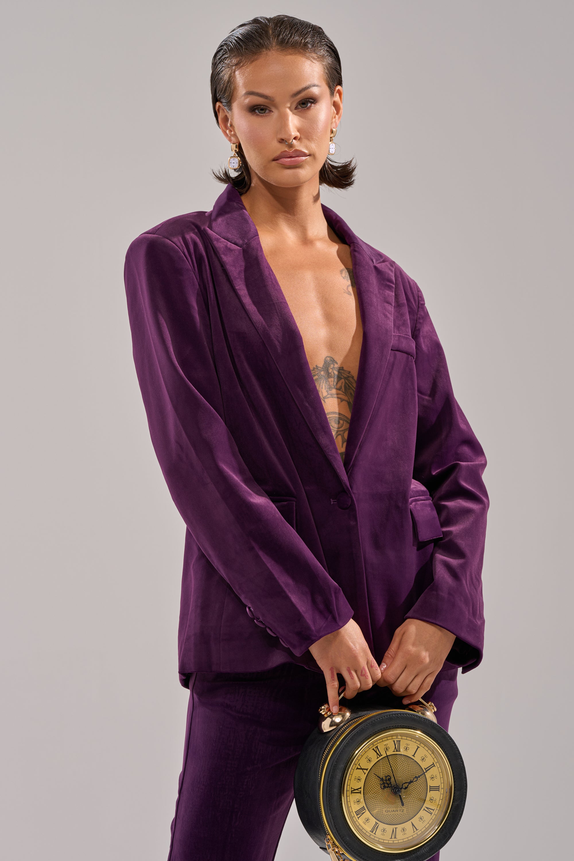 Wearing the I ADORE YOU BLAZER in purple velvet, a confident person with short slicked-back hair, statement earrings, and chest tattoos holds a vintage-style clock against a plain neutral background.