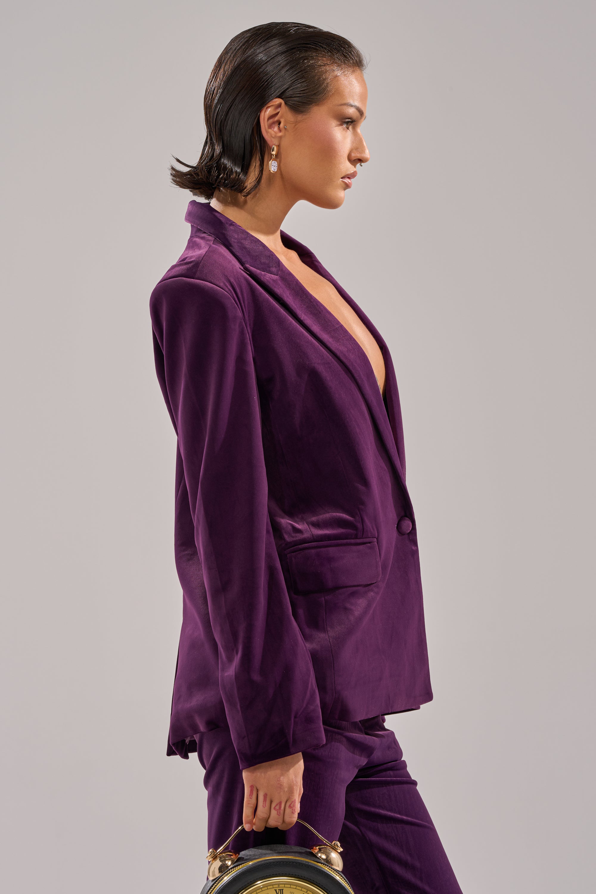 A woman with slicked-back hair wears the I ADORE YOU BLAZER in deep purple velvet and holds a small round handbag, facing left against a plain light gray background.