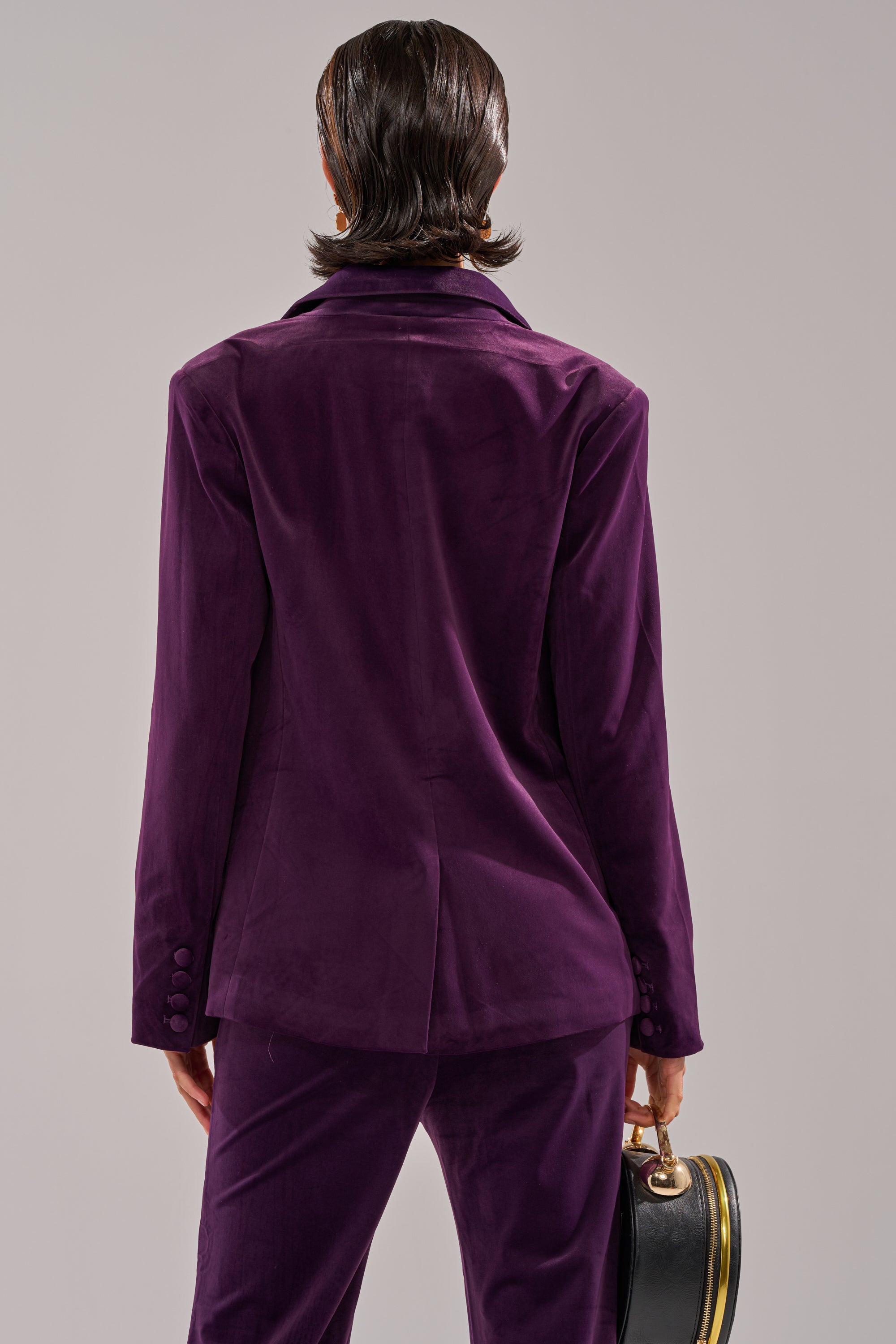A person with short dark hair, seen from behind, wears the I ADORE YOU BLAZER in deep purple velvet and holds a black round handbag with a gold clasp against a plain light-colored background.