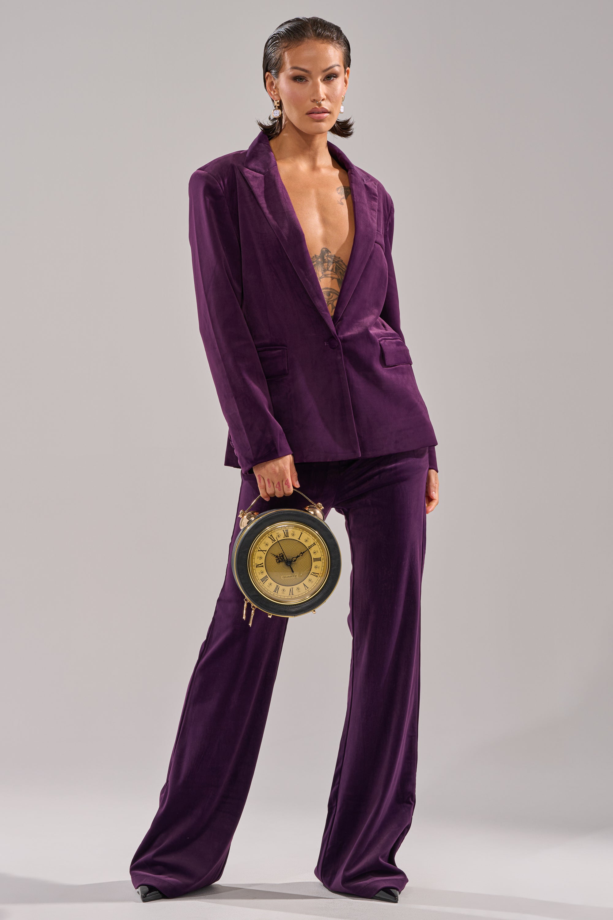 Wearing the I ADORE YOU BLAZER in deep purple velvet with no shirt underneath, a confident individual poses holding a round clock like a handbag against a plain light gray background.