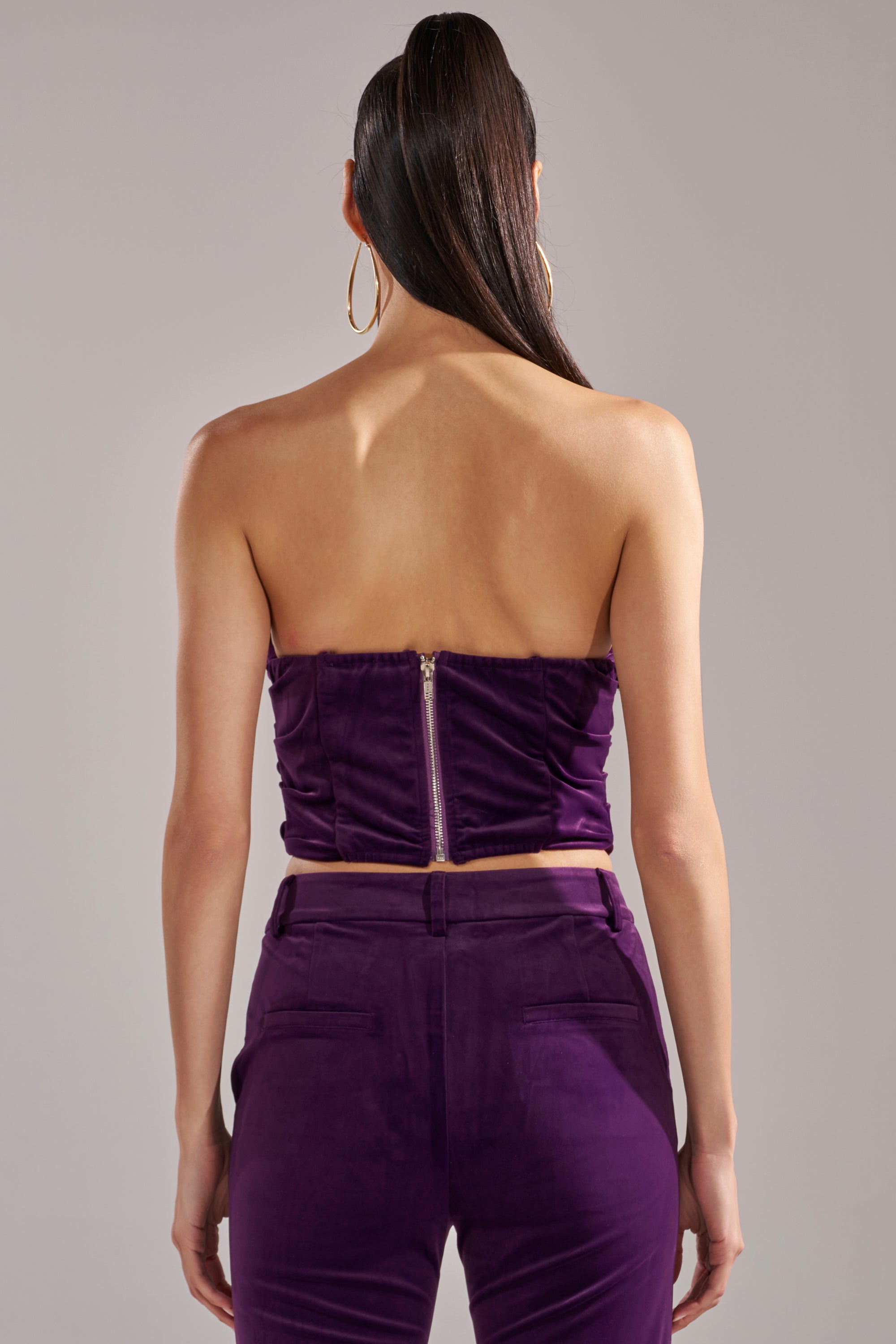 A person with long dark hair in a high ponytail is seen from the back, wearing the I ADORE YOU CORSET in eggplant purple with a visible zipper, matching high-waisted velvet pants, and large hoop earrings.