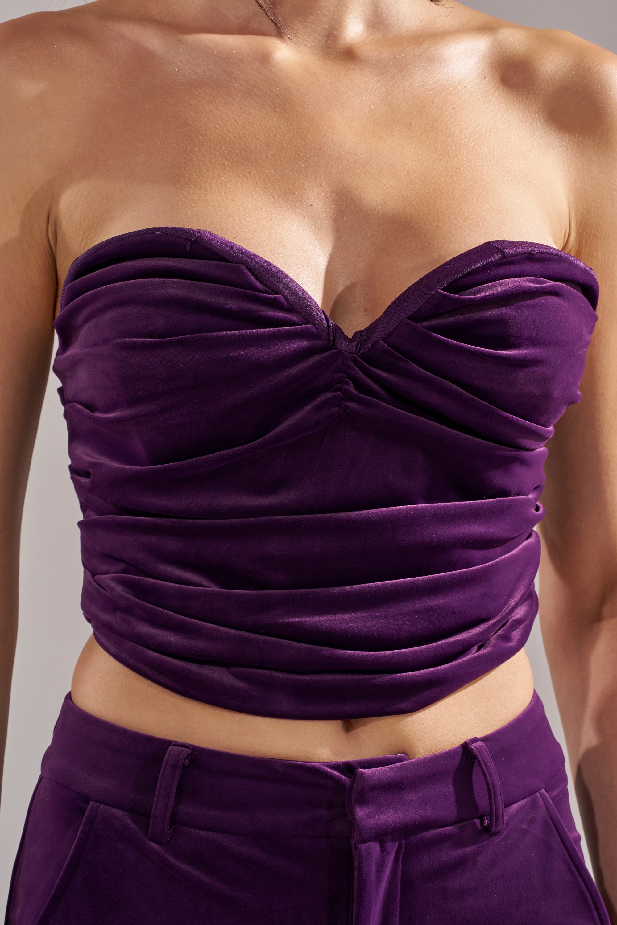 A close-up of a person wearing the I ADORE YOU CORSET, an eggplant purple velvet top with gathered details and a sweetheart neckline, paired with matching pants. The photo is cropped at the shoulders and hips.