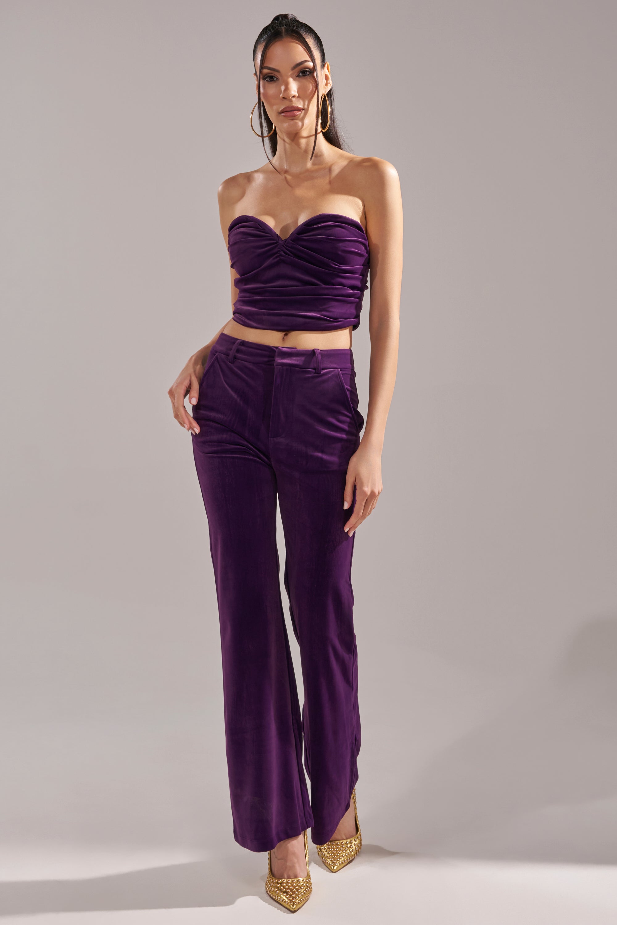 A woman poses in a studio wearing the I ADORE YOU CORSET, an eggplant purple, ruched strapless top, paired with matching high-waisted wide-leg pants, gold pointed-toe heels, and hoop earrings against a light gray background.