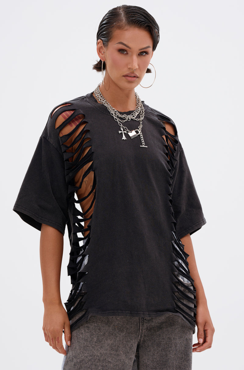 A woman wears the SHAINA DISTRESSED OVERSIZED TEE featuring large side and sleeve slits, paired with layered necklaces, hoop earrings, and grey jeans, standing against a plain white background. 0